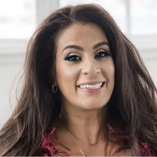 A woman with long dark hair, brown eyes, and brown skin smiles at the camera. She is in front of a white wall with windows.